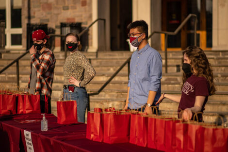 Four masked students stand behind a table, covered in a red table cloth. Red paper bags are on top of the table.