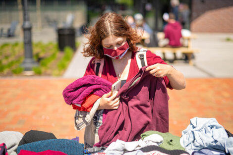 A student wearing a masks looks closer at a sweater, while holding several others in front of a table of clothing at the pop-up thrift shop.