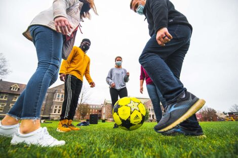 Five masked students stand in a circle, kicking a ball in front of Hogg Hall and Farinon College Center