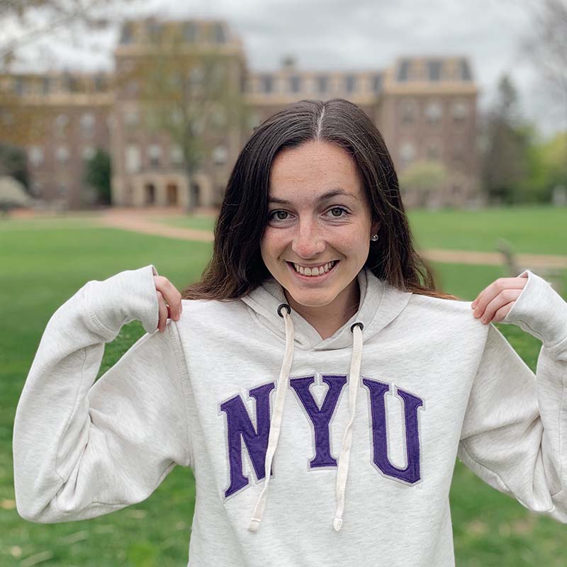 Clare Mengel stands in front of Pardee Hall in a white hoodie with NYU in purple letters