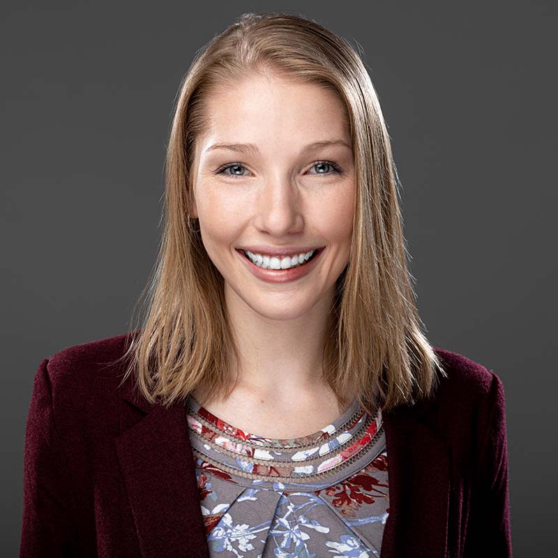 Sasha Neefe smiles in front of a gray backdrop. She wears a maroon blazer.