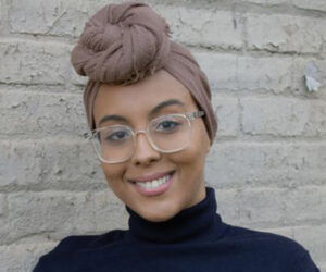 Imane Halal smiles, wearing glasses and a pink headwrap
