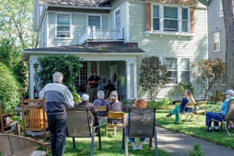 performer sings and plays guitar on a porch with audience in folding chairs