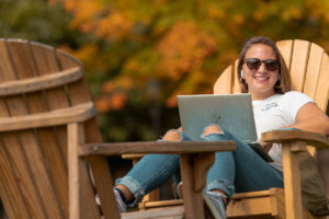 student wearing sunglasses and holding laptop on lap smiles sitting in Adirondack chair on Quad