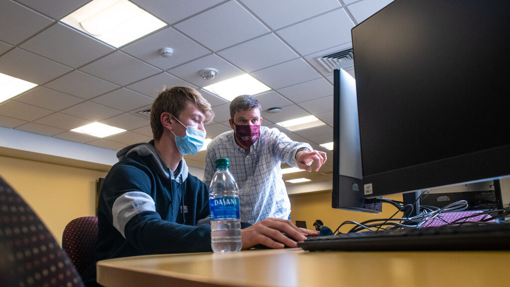 Trent Gaugler wears Lafayette mask, points at a computer screen with a student in a tech lab