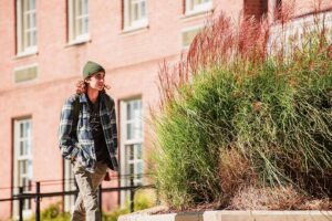 student in winter hat and flannel shirt walks past brick building on campus