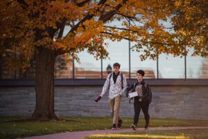 two students walk on campus, Skillman and orange foliage behind them