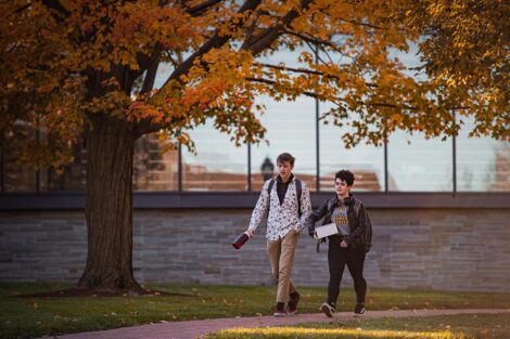 two students walk on campus, Skillman and orange foliage behind them