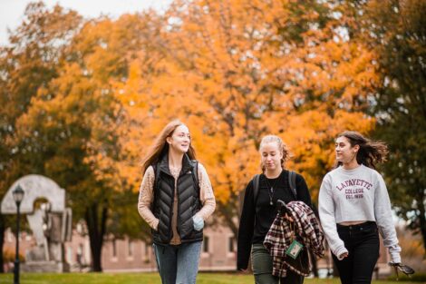 three students walk and talk outdoors on campus, orange foliage is behind them
