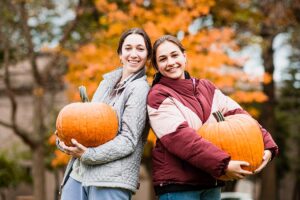 two students smile while holding pumpkins, orange foliage is behind them