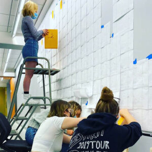 student stands on a ladder, others sit on the floor to attach pieces of paper to a wall