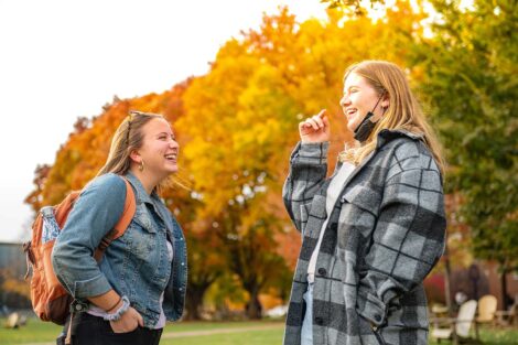 two students smile and laugh while talking on the Quad, golden foliage is behind them