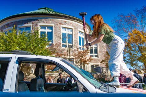 A student stands on top of an SUV and hits it with a sledgehammer.