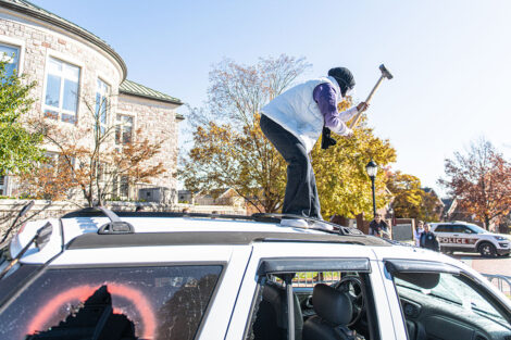 A student stands on top of an SUV and hits it with a sledgehammer.