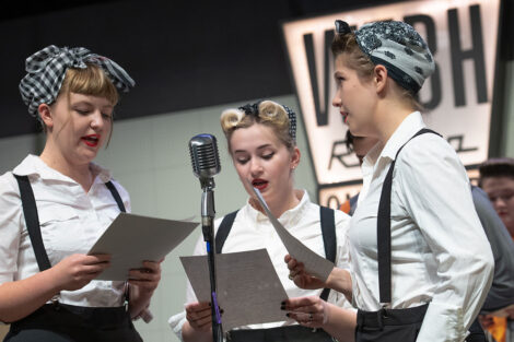 student performers in vintage dress gather on stage with vintage microphones