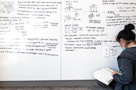 student sits on the ground and looks in a book with a giant white board covered in equations