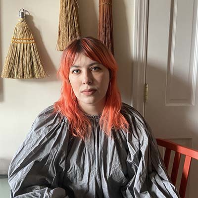 Krystal DiFronzo with orange hair sits before a wall of brooms