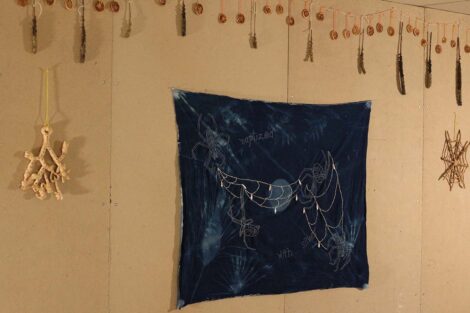 Blue tapestry hangs on wall beneath a string of feathers and dried lemons. On either side of the tapestry is a bread sculpture