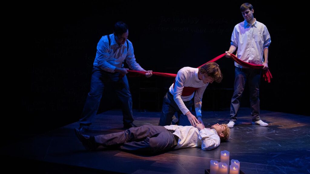 Two men hold a long piece of red fabric which is wrapped around another man who looks at a man lying dead on the floor