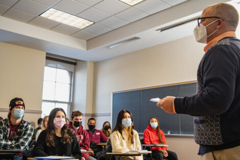 Students wearing masks, sit inside of a classroom while Brett Hendrickson speaks.