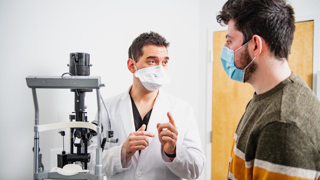 Doctor stands alongside eye exam equipment and speaks with student