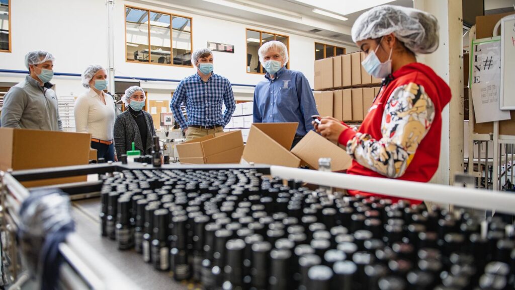 Group of people stand at end of assembly line where bottles are gathering