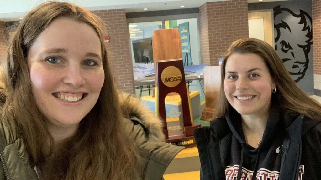 Host and extern stand in front of NDSU NCAA Championship trophy