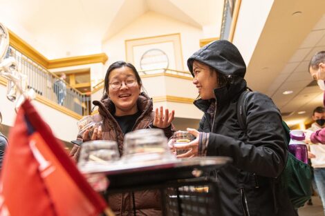 two students in winter coats smile while taking cupcakes from a table