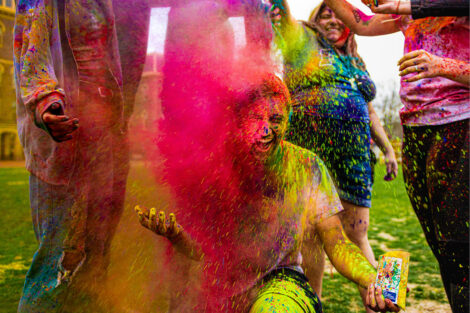 A student sits as other students pour color powder in the air, landing on top of them.
