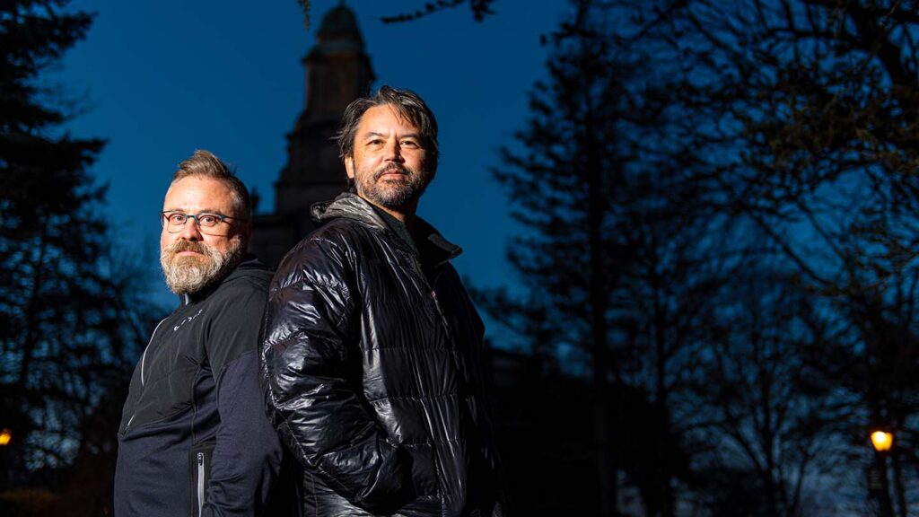 Profs. Robert Blunt and Christopher Lee stand back to back in front of Colton Chapel at dusk