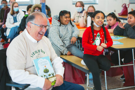 Sal Panto laughs while holding a book and sitting with elementary school students