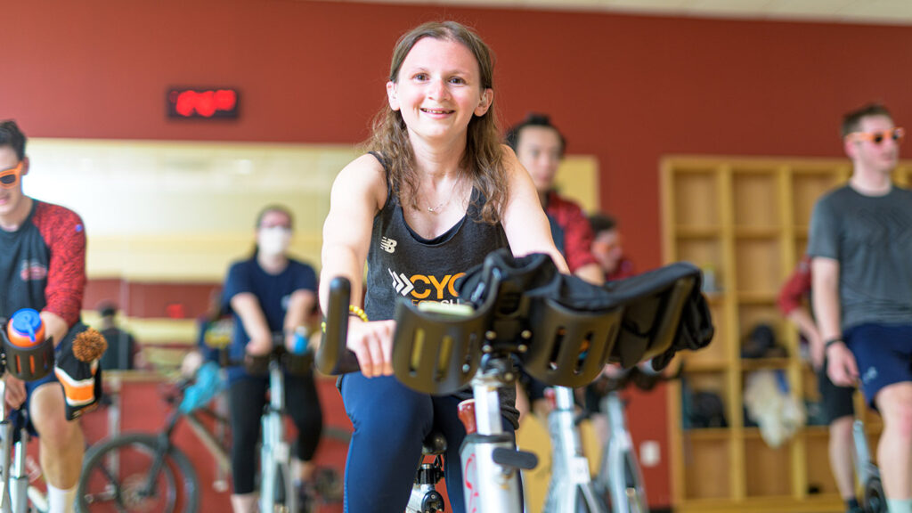 Perry Zimmerman smiles as she rides a spin bike, other students on spin bikes behind her