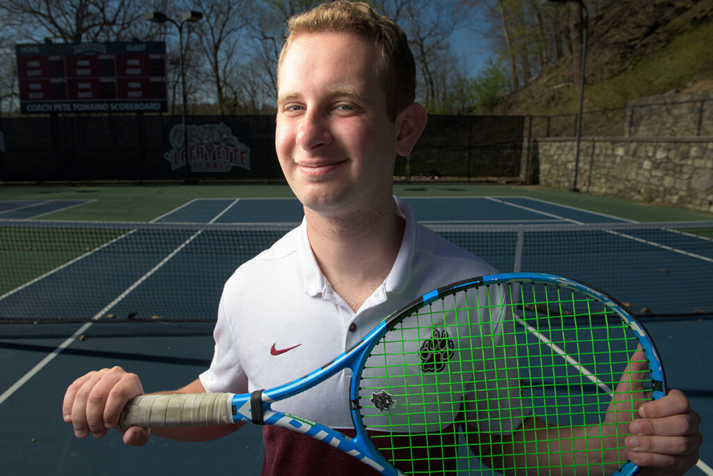 Ross Coleman smiles while holding a tennis racket at a tennis court.