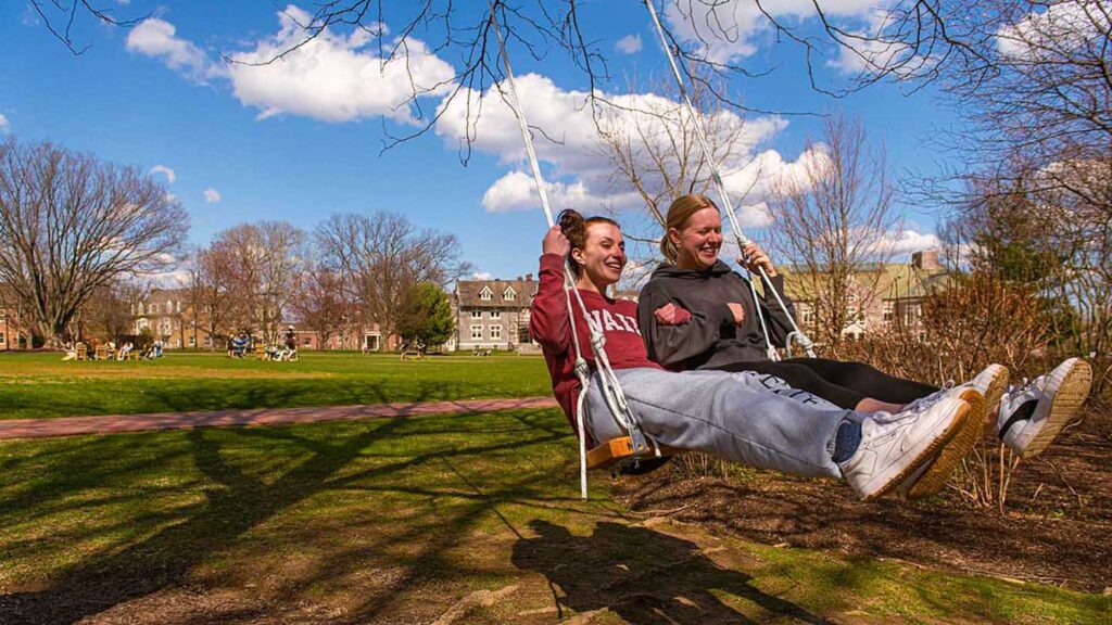 two students sit on a swing and smile, blue sky behind them