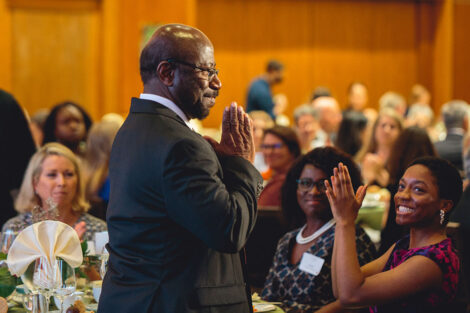 Prof. Rex Ahene stands with his hands together in a prayer gesture and smiles as colleagues sitting at tables applaud
