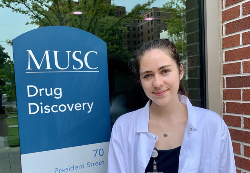 Camille Carthy wears a lab coat and stands outside a building with a sign that reads MUSC drug discovery