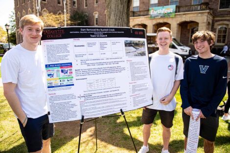 three students stand next to a poster highlighting research out on the Quad