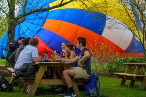 students sit together at a picnic table on the Quad as the hot air balloon is lying on its side on the Quad
