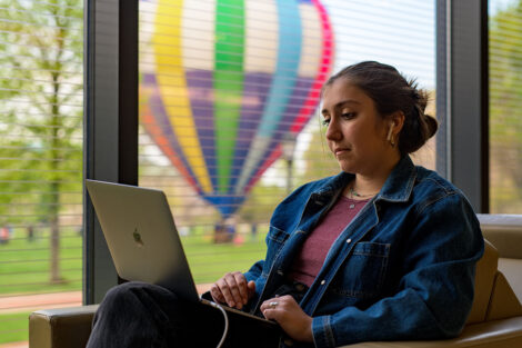 student works on a laptop in Skillman, a colorful hot air balloon is visible through a window