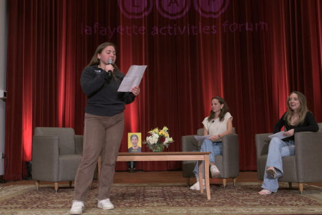 student addresses audience on stage in Colton Chapel using microphone, two students sit on stage