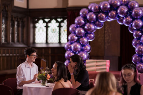 students stand at high tables in Hogg Hall, purple balloon arch in background