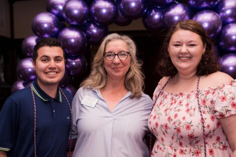 Amy Blythe stands and smiles with two students in front of purple balloon arch