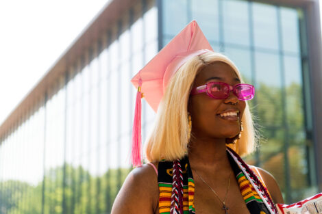 A student wears a pink graduation cap and matching sunglasses, smiling.