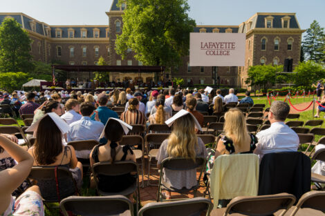 crowd of family members sit in folded chairs arranged in rows on the Quad, stage and big screen with Lafayette College logo in distance