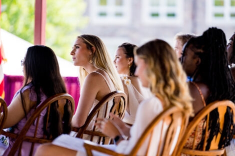 students in dresses sit in chairs on stage, Hogg Hall in background