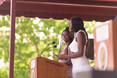 students in dresses stand a podium and speak