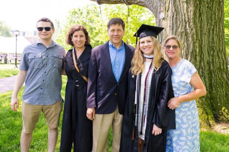 family members smile with a graduate in cap and gown in front of a tree on the Quad