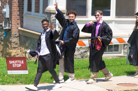 students in gowns hold caps in hands as they walk down a sidewalk