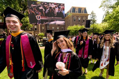 students in caps and gowns smile as they walk across the Quad, Pardee in the background