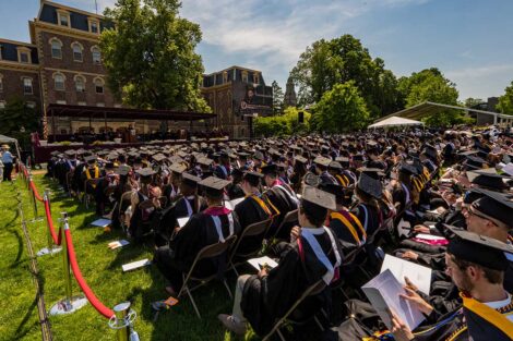 graduates in caps and gowns sit on the Quad in chairs arranged in rows and face a stage in front of Pardee